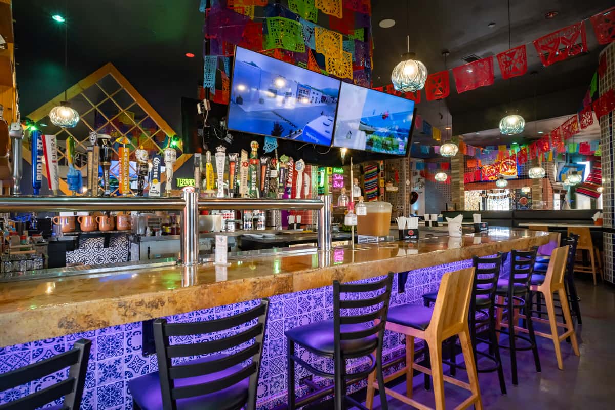 Vibrant bar area decorated with colorful papel picado, multiple beer taps, and large TVs above a tiled counter.