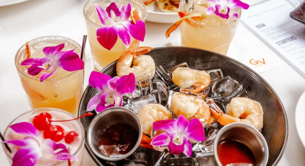 Shrimp cocktail platter with orchids and tropical drinks on ice at an outdoor garden restaurant table, highlighting sensory dining experience.