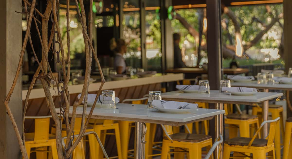 Outdoor restaurant patio with yellow metal chairs, white tabletops, rolled napkins, and greenery creating a relaxed garden dining atmosphere.