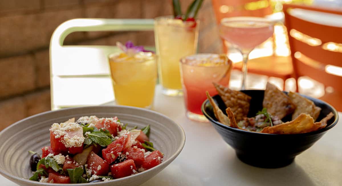 Watermelon and feta salad with fresh mint alongside tortilla chips and colorful cocktails on a sunlit patio table.