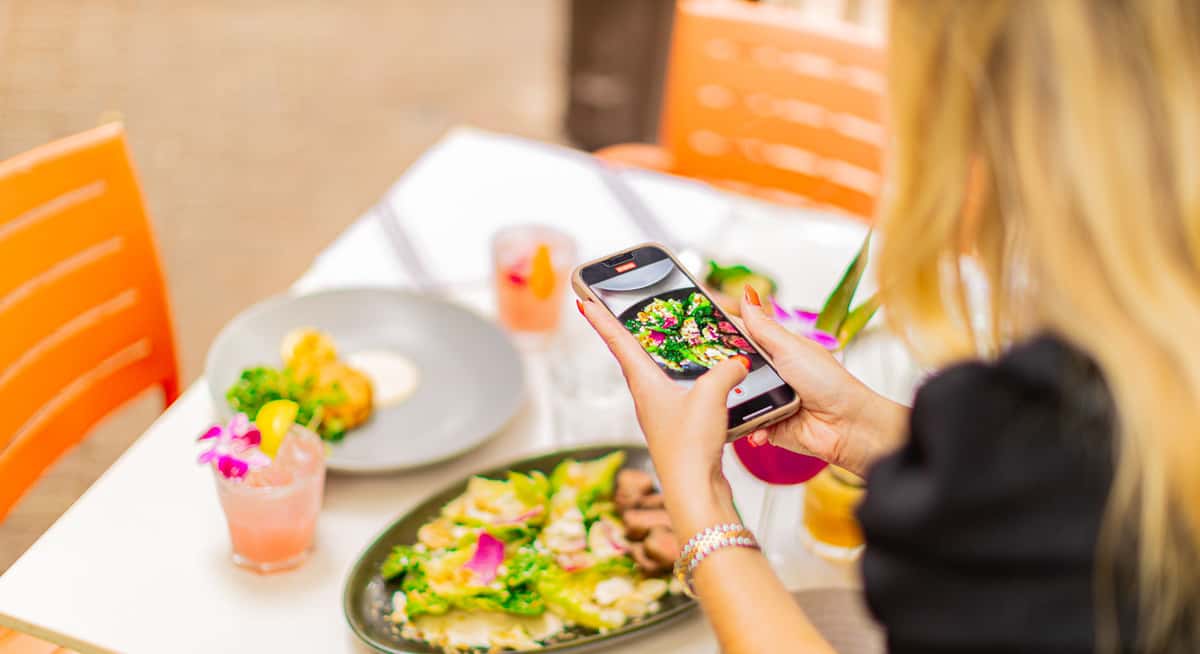 Guest taking photo of colorful plated salad and cocktails with smartphone at outdoor restaurant table.