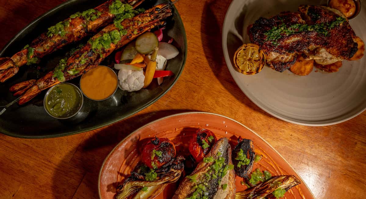 Grilled jumbo prawns with green herb sauce, roasted fish with charred tomatoes and fennel, and crispy-skinned chicken with grilled lemon on a wooden table.