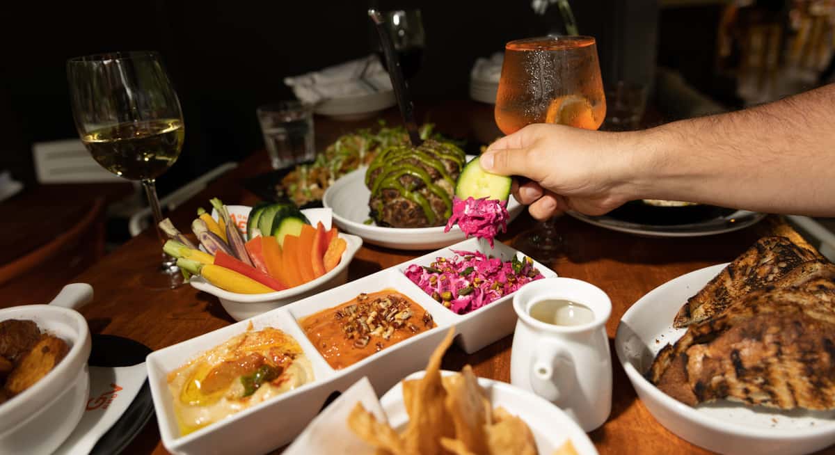 Assorted shared plates with hummus, dips, fresh vegetables, grilled bread, and wine served at a warmly lit restaurant table.