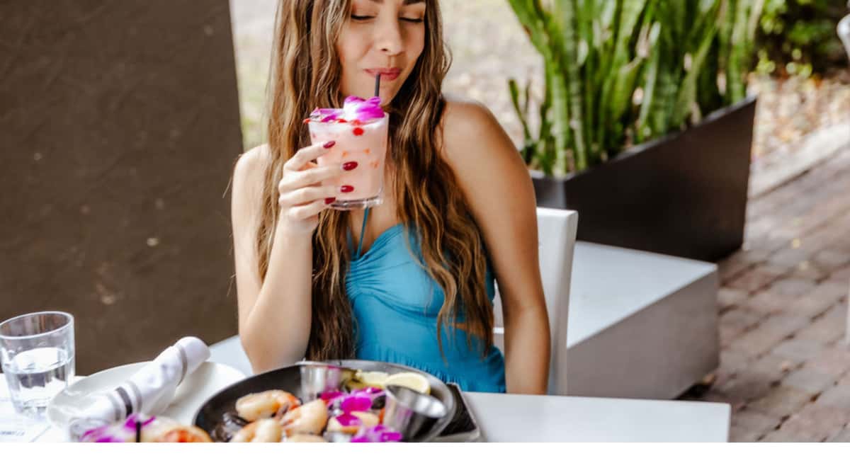 Woman in blue dress sipping pink tropical cocktail with orchid garnish at outdoor restaurant table with seafood dish.