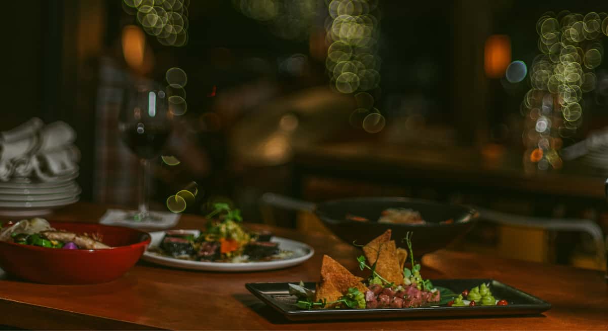 Evening restaurant table with wine, small plates, and appetizers under warm string lights creating cozy ambiance.