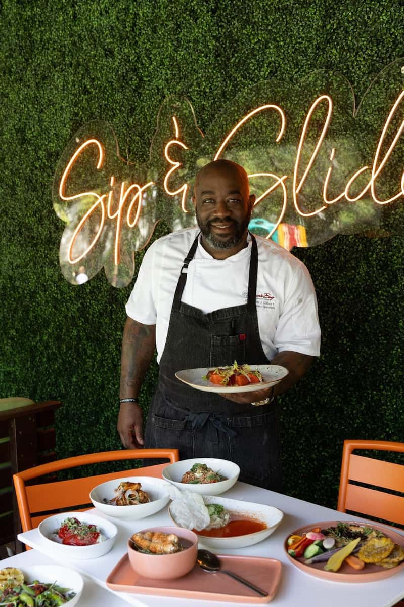 Chef presenting plated dishes in front of a glowing restaurant sign and greenery wall