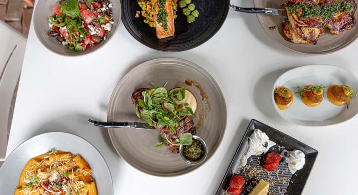 Overhead view of assorted restaurant dishes including steak with greens, grilled salmon, pasta, salad, croquettes, and dessert on a white table.