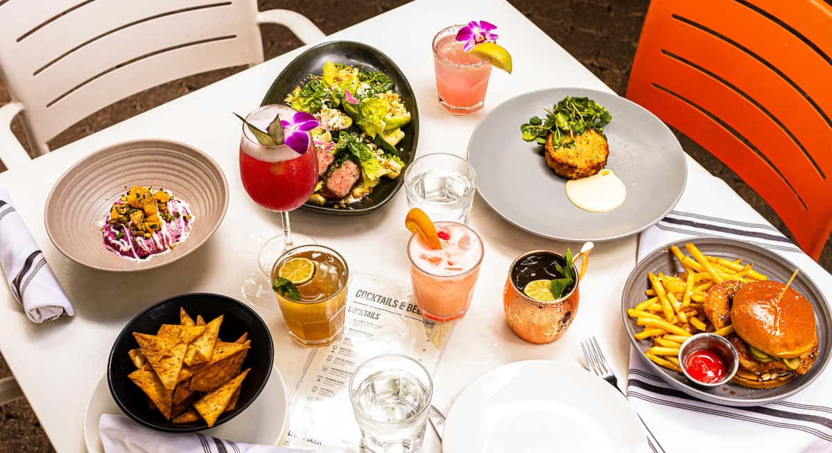 Overhead view of restaurant table with cocktails, salad, burger and fries, chips, and assorted appetizers.