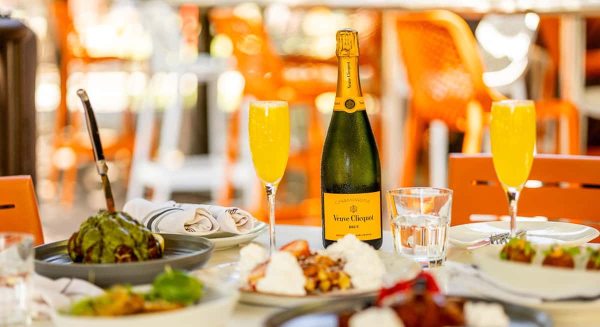 Bottle of Veuve Clicquot champagne with two mimosas on an outdoor table surrounded by brunch plates and orange patio chairs.