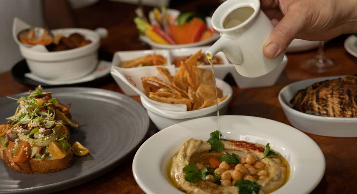 Mediterranean appetizer spread with hummus, olive oil drizzle, grilled bread, dips, fresh vegetables, and grilled protein on wooden table.