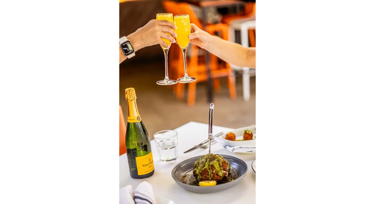 Two people toasting mimosas above a table with champagne bottle and plated appetizer in an outdoor restaurant setting.