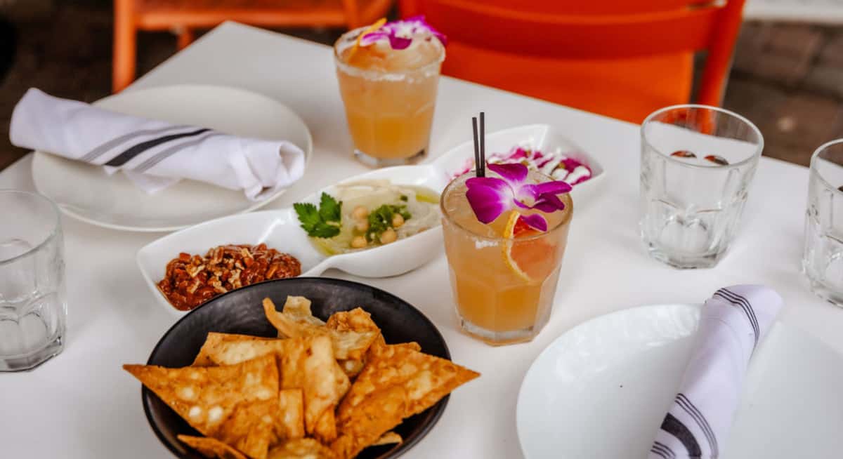 Garden restaurant table with cocktails topped with orchids, crispy chips, and Mediterranean-style dips in an open-air setting.