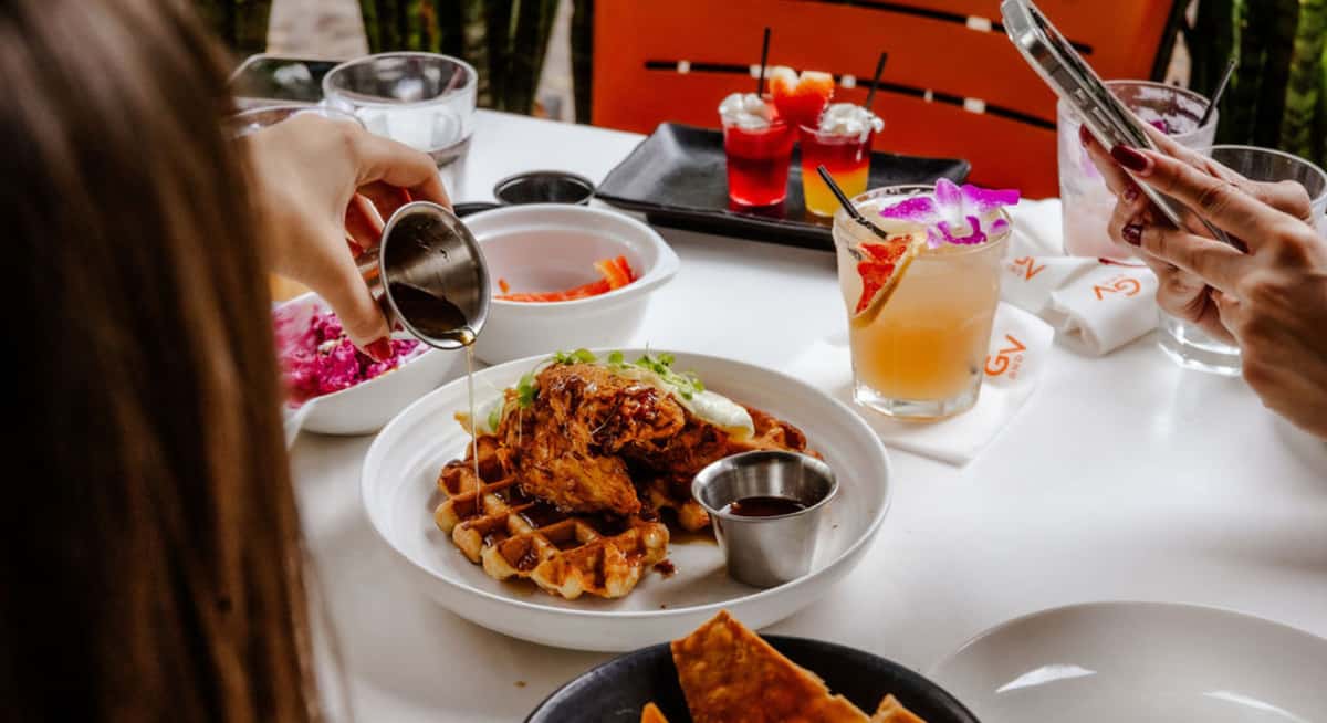 Chicken and waffles brunch plate with syrup being poured, tropical cocktail garnished with orchid, and guests dining outdoors.