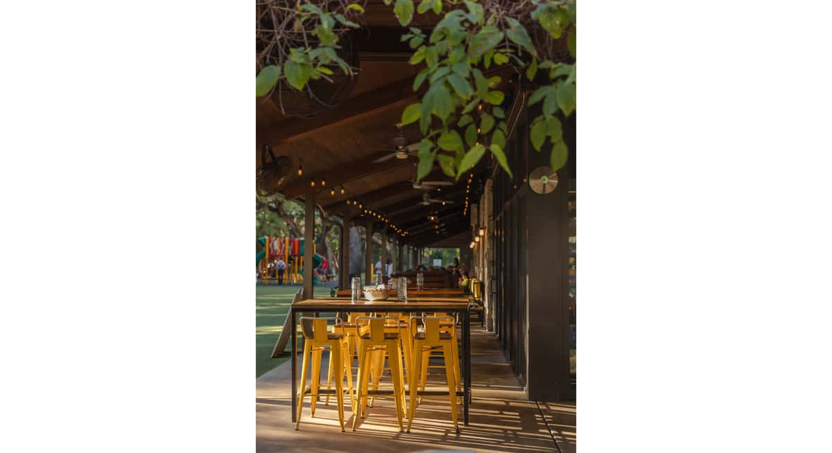 Covered outdoor restaurant seating with yellow bar stools, wooden tables, string lights, and park playground in the background.