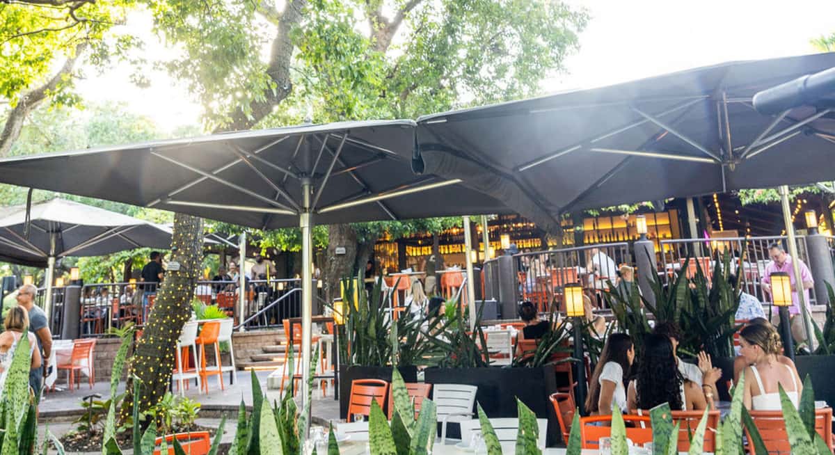 Busy outdoor restaurant patio with large black umbrellas, orange chairs, and guests dining in a lush garden setting.
