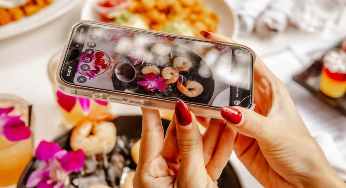 Guest taking photo of shrimp dish with orchid garnish using smartphone at outdoor restaurant table.