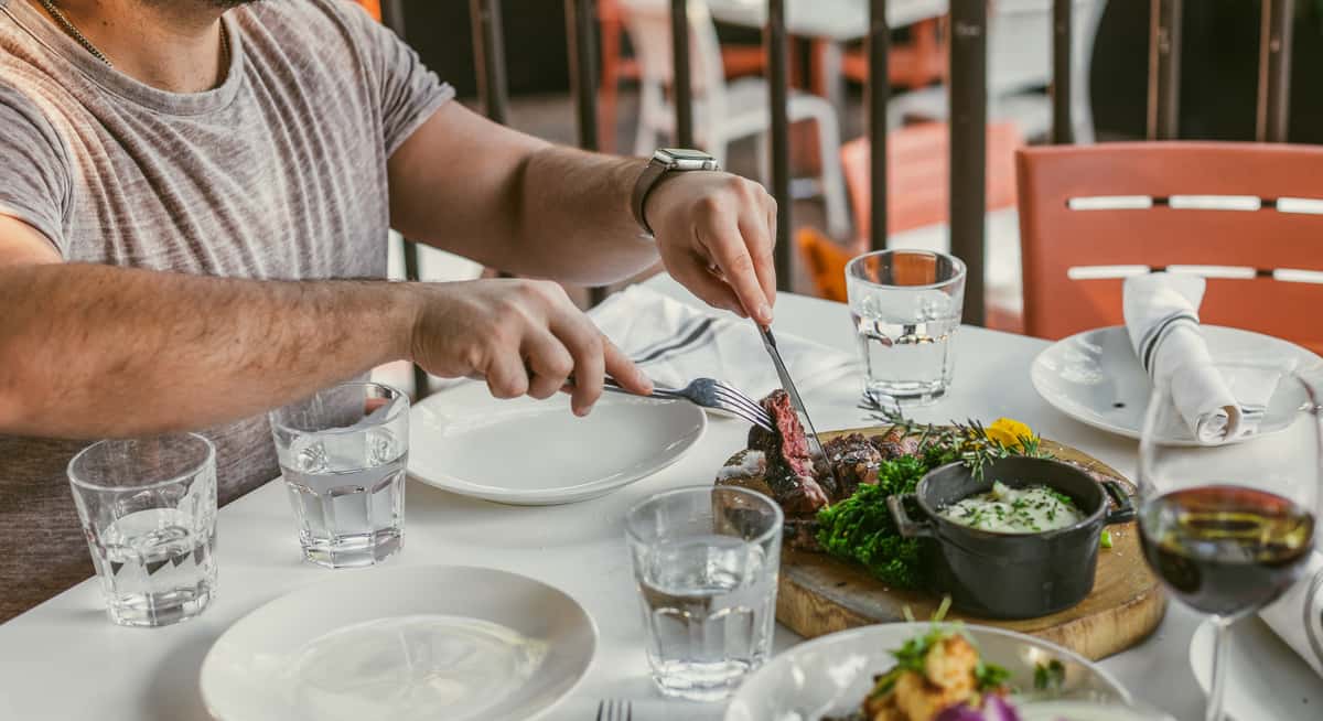 Man cutting medium-rare steak at outdoor restaurant table with wine, water glasses, and side dishes.