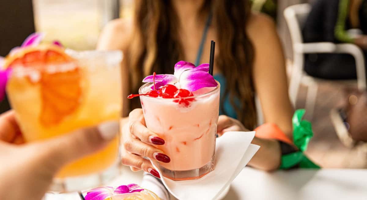 Close-up of tropical cocktails garnished with orchids and cherries as two guests toast at an outdoor restaurant table.