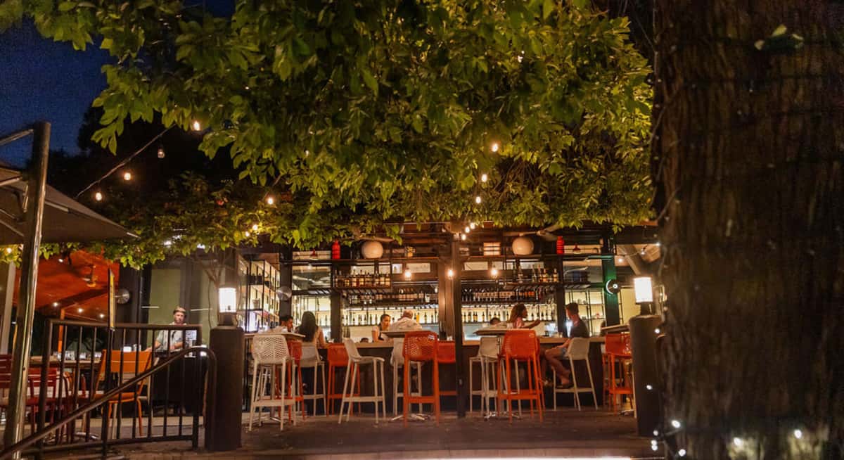 Open-air restaurant bar at night with orange and white bar stools, string lights, and guests seated under leafy trees.