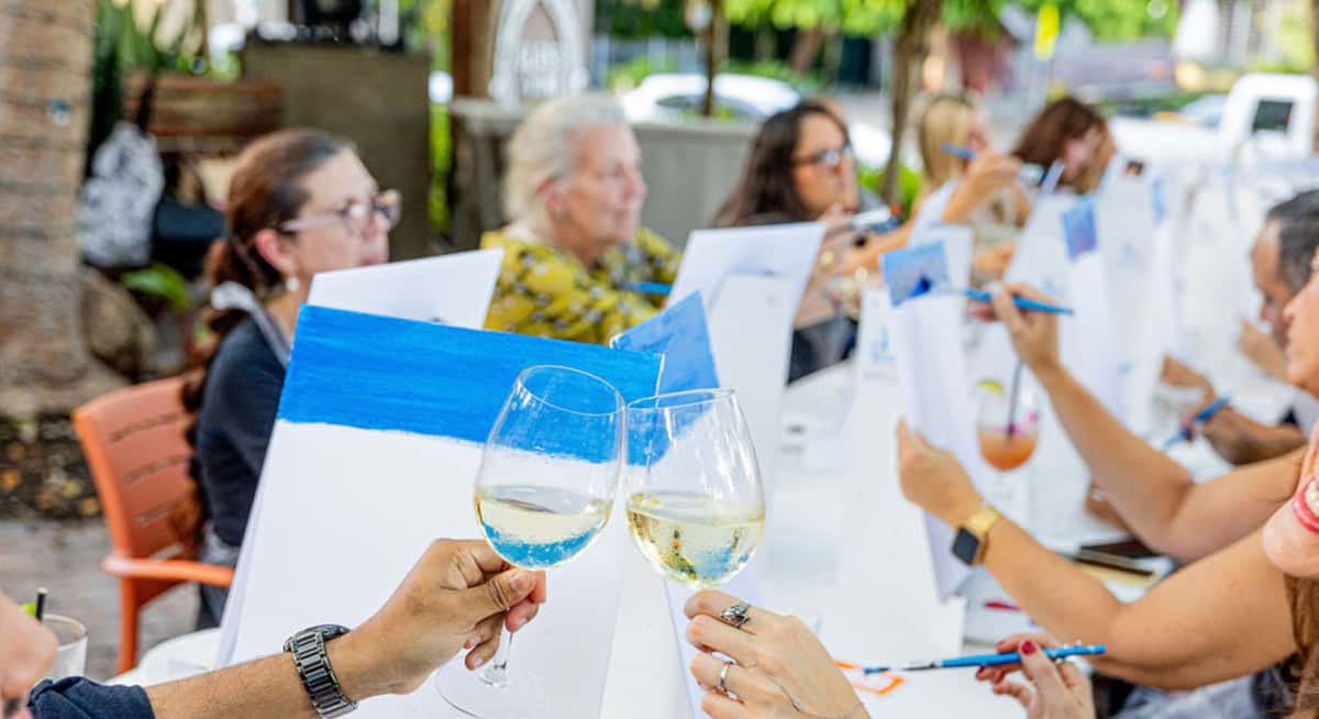 Group of friends enjoying wine and painting together at an outdoor garden dining event, toasting glasses during a social gathering.