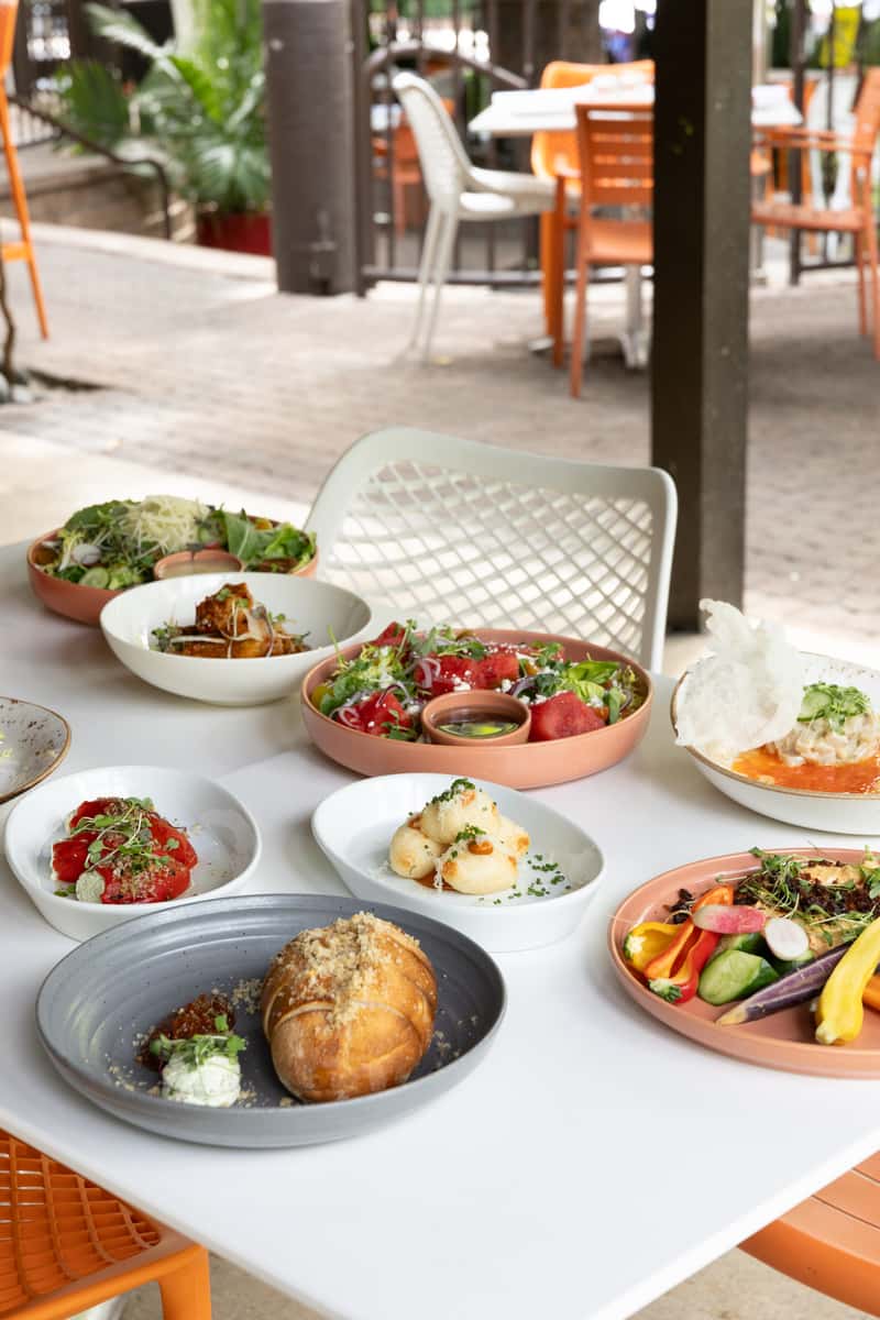 Outdoor table spread with salads, small plates, bread, vegetables, and plated dishes on a restaurant patio