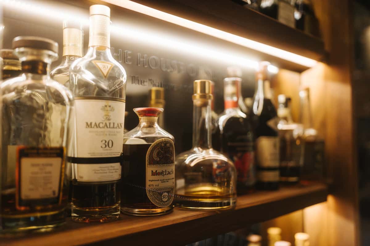 Assorted liquor bottles lined up on a shelf in a dimly lit bar setting.