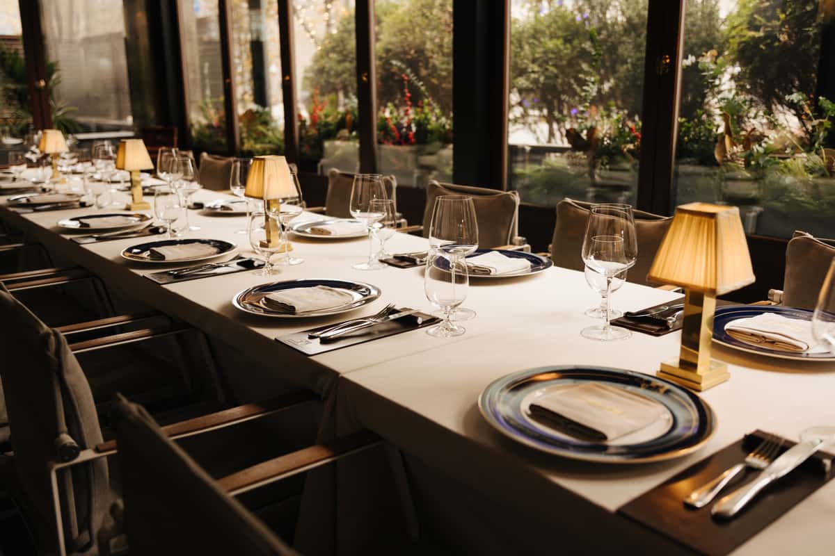 Long dining table set with plates and wine glasses near large windows.