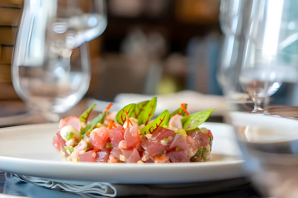 Tuna tartare plated on a white dish with garnish, with wine glasses blurred in the background.