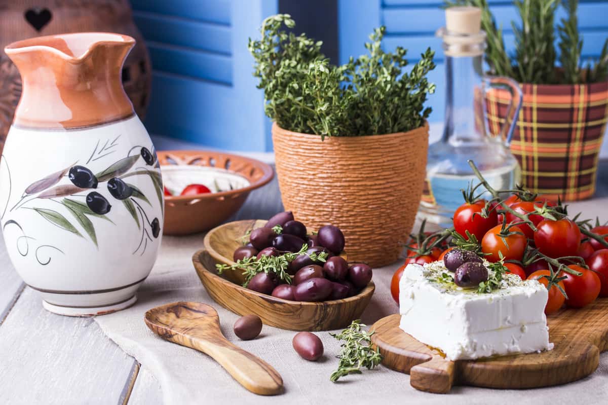 Fresh Greek ingredients including feta cheese topped with olives and herbs, cherry tomatoes on the vine, and a bowl of Kalamata olives.