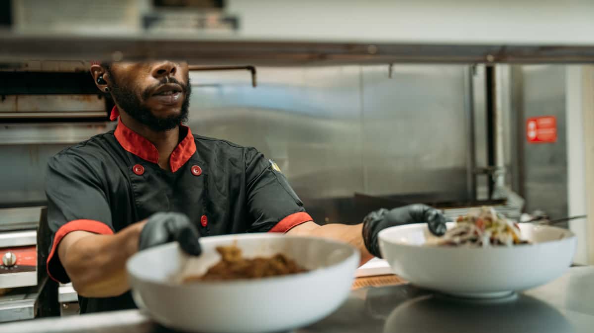 Chef in black and red uniform plating freshly prepared dishes in a commercial kitchen, ready for service.