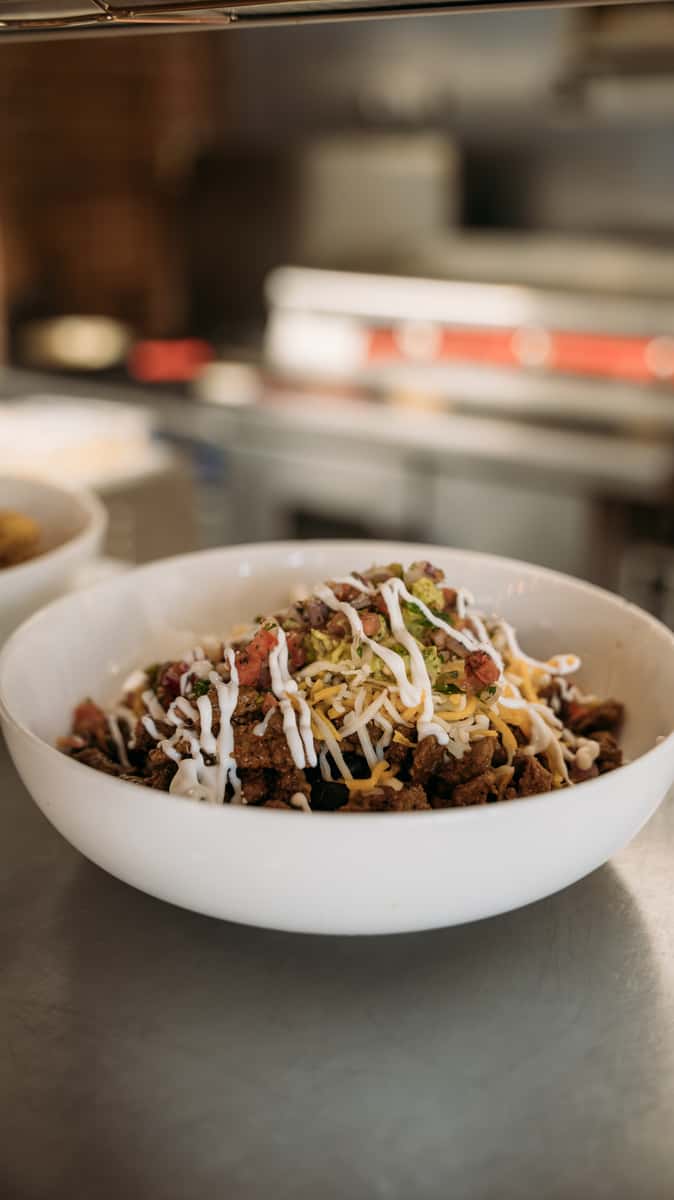 Freshly prepared burrito bowl with seasoned beef, shredded cheese, pico de gallo, and a drizzle of sour cream, served in a white bowl at a restaurant kitchen counter.