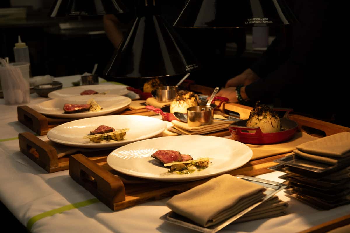 Steak dishes being plated under heat lamps in a steakhouse kitchen.