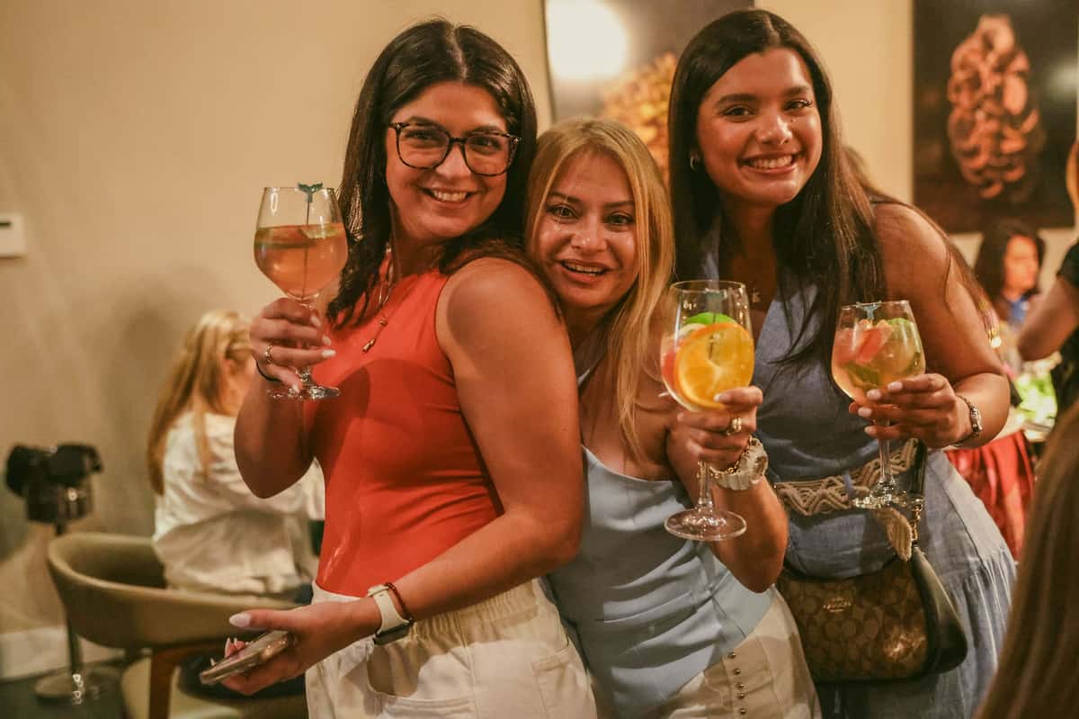 Three smiling guests raise fruit-garnished cocktails while celebrating together at an indoor private party.