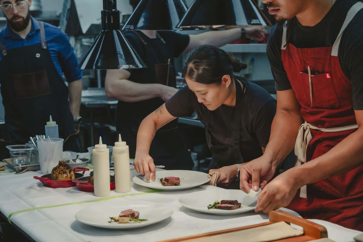 Chefs plating sliced steak dishes in a restaurant kitchen.