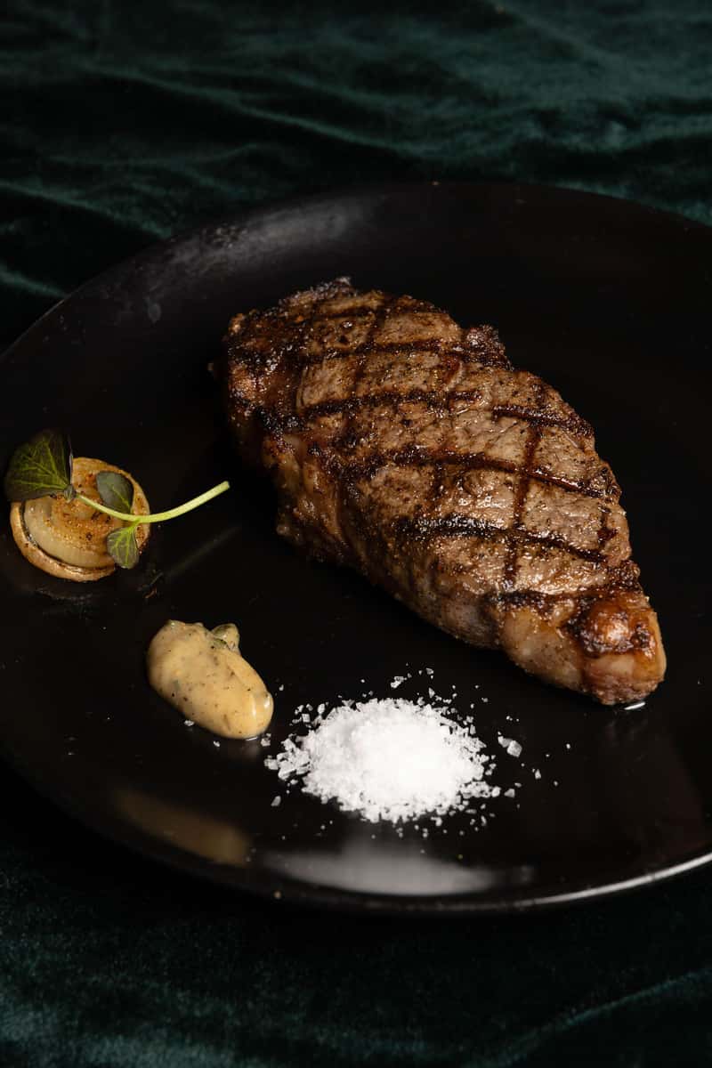 Grilled steak with char marks served with sea salt, sauce, and roasted onion on a black plate.