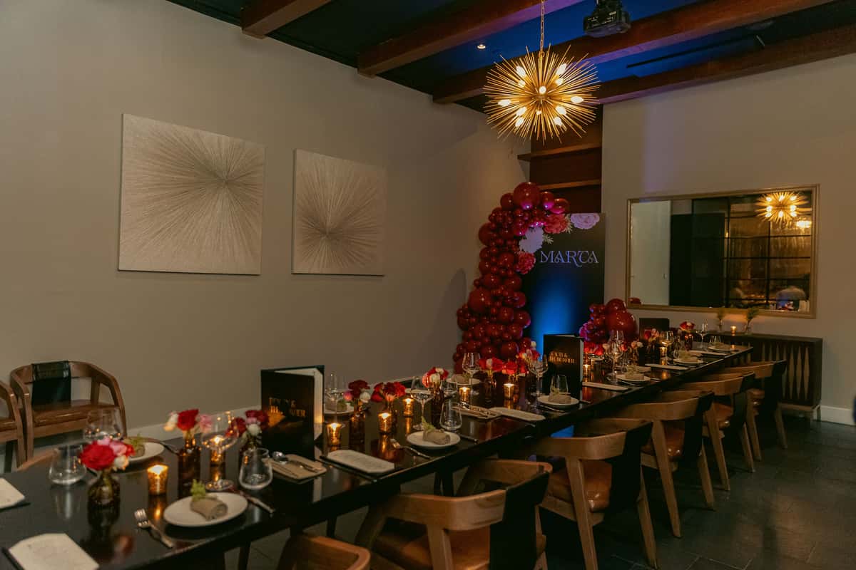 An elegant modern steakhouse private dining room features a long table with floral centerpieces, candlelight, contemporary lighting, and red balloon decorations.