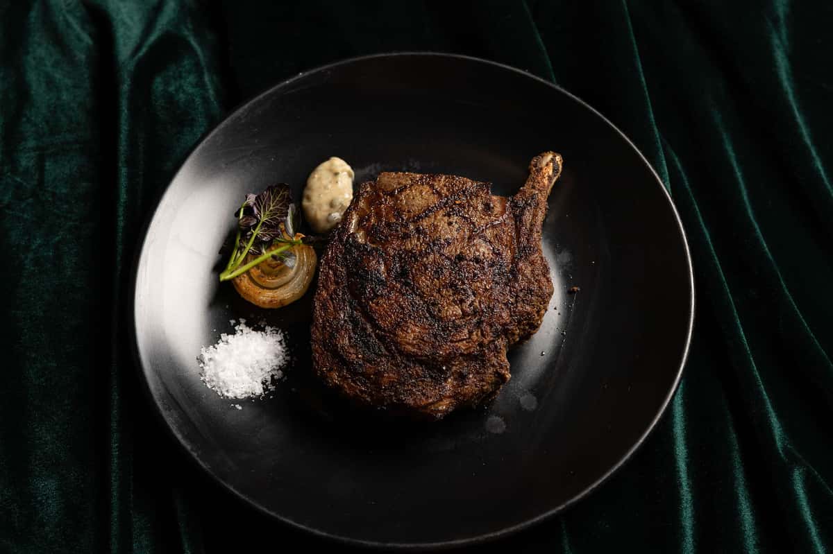 Grilled dry-aged cowboy steak served with salt, sauce, and onion garnish.