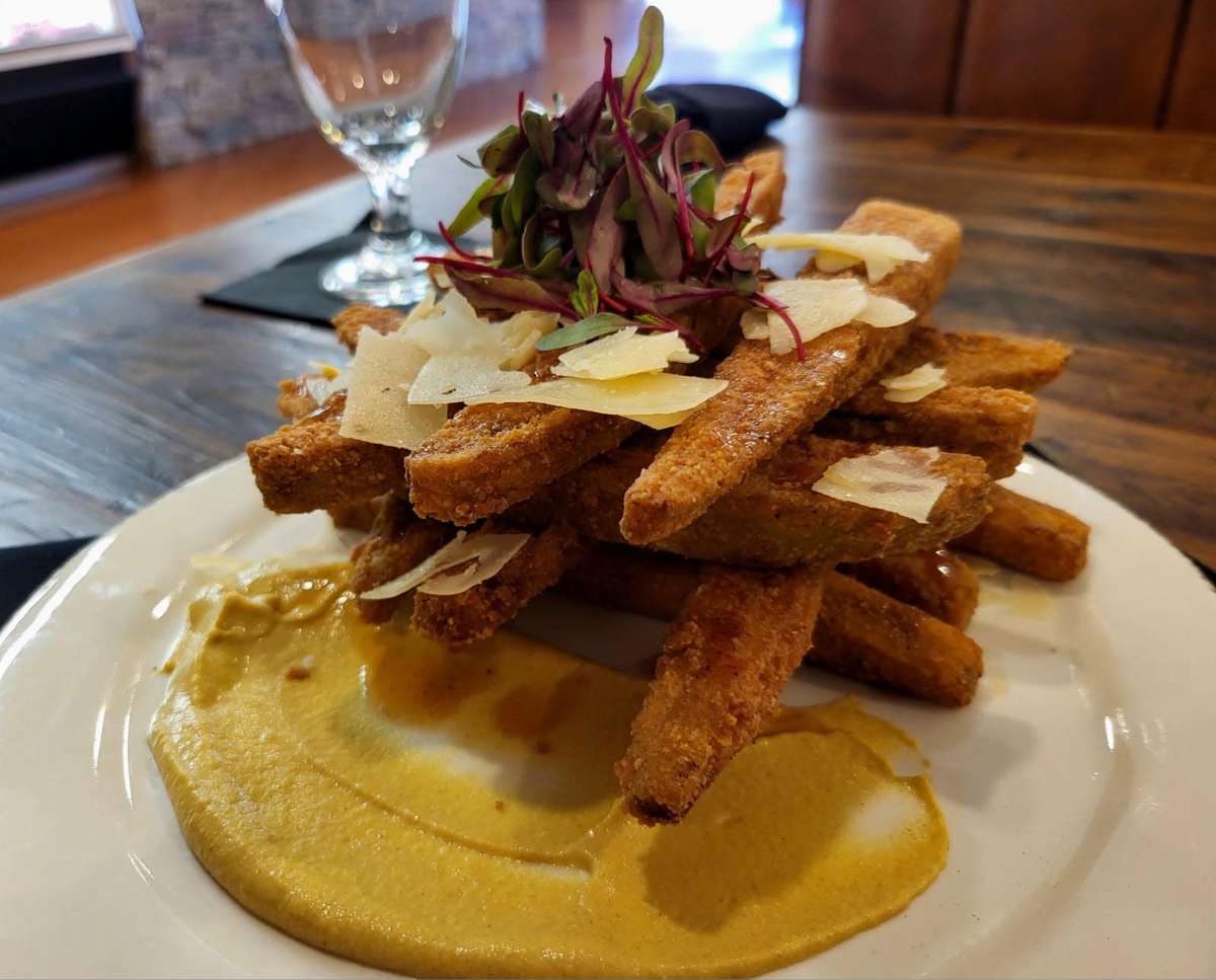 Crispy fried polenta fries stacked neatly, topped with shaved Parmesan and microgreens, served with a generous smear of creamy mustard dipping sauce.