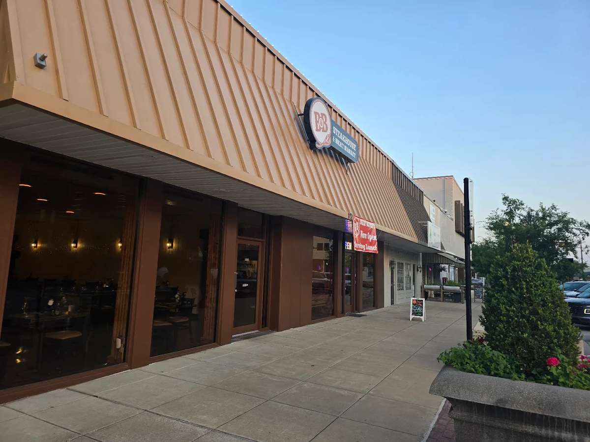Street view of B&B Steakhouse & Meat Market with large front windows, tan metal awning, and a banner announcing it's now open.