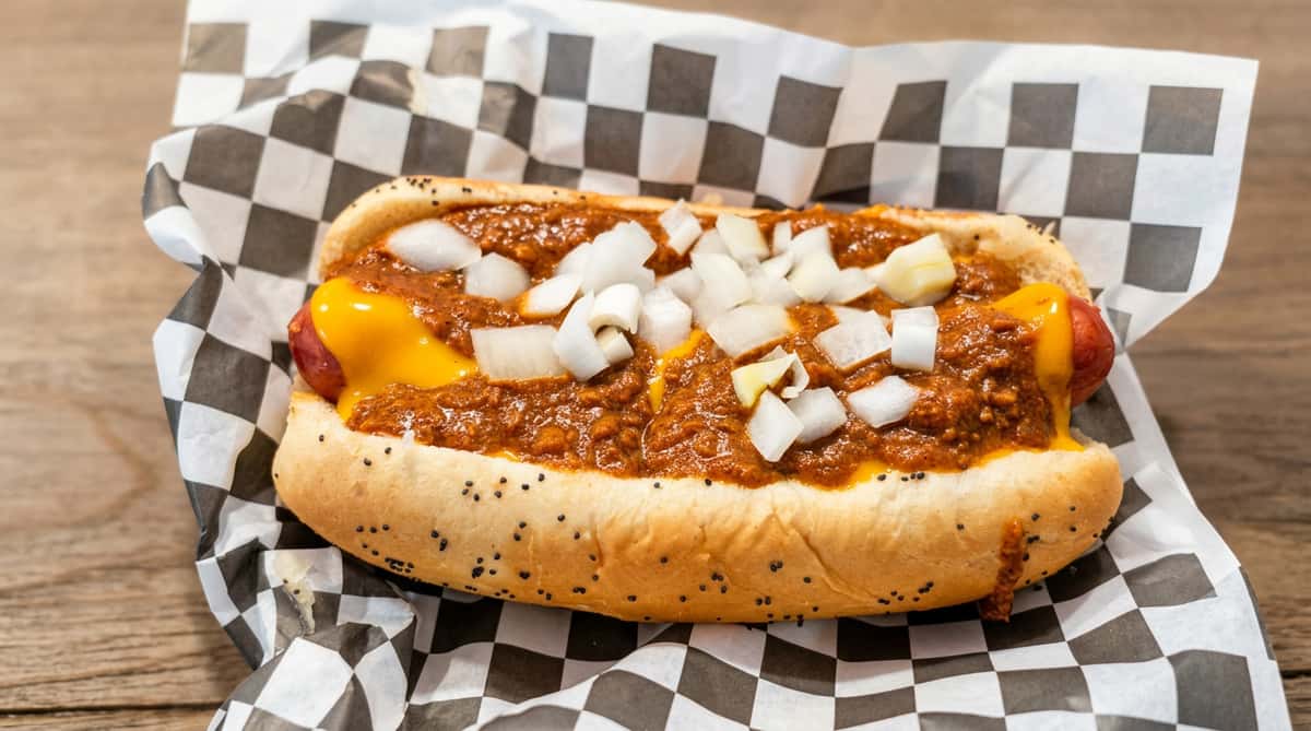 Chili cheesedog in a poppy seed bun, topped with diced white onions, on checkered paper.