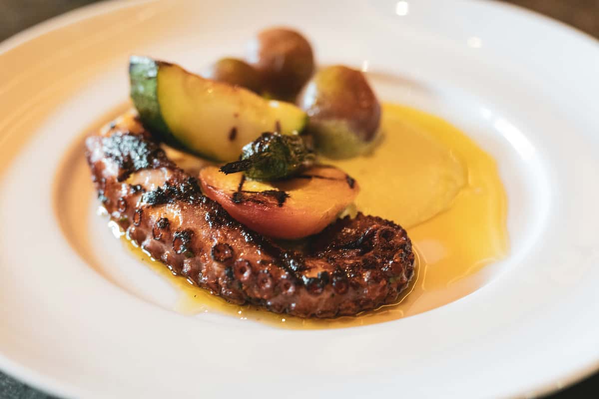 Grilled octopus tentacle served with roasted zucchini, seared peach, fingerling potatoes, and a golden olive oil emulsion on a white plate.