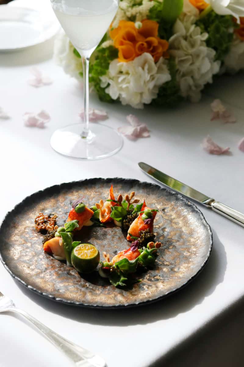 Elegant seafood tasting plate with lobster medallions, black caviar, citrus, and edible greens, paired with a clear cocktail on a white-linen table.