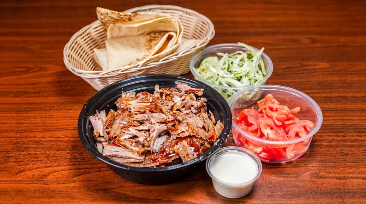 Shawarma plate with sliced meat in a black bowl, pita bread in a basket, diced tomatoes, pickled onions, and white sauce.