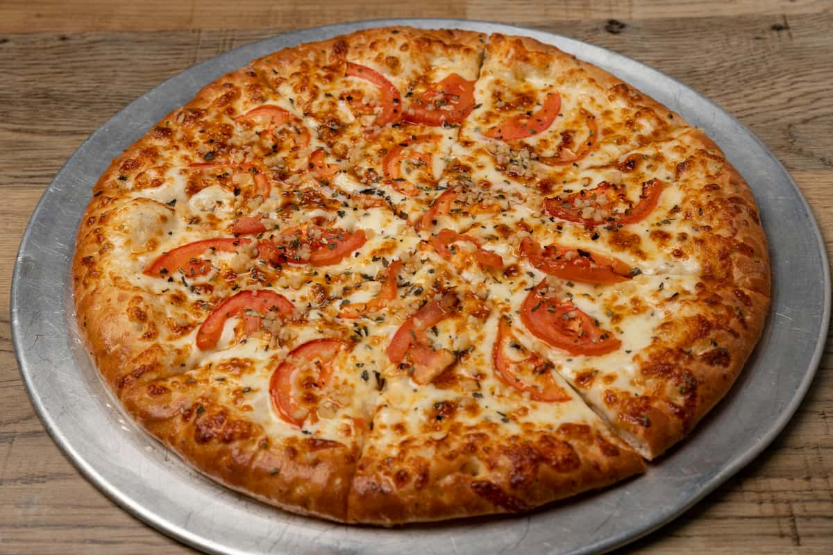 Cheese pizza topped with fresh tomato slices, minced garlic, and herbs, served on a metal tray over a wooden table.