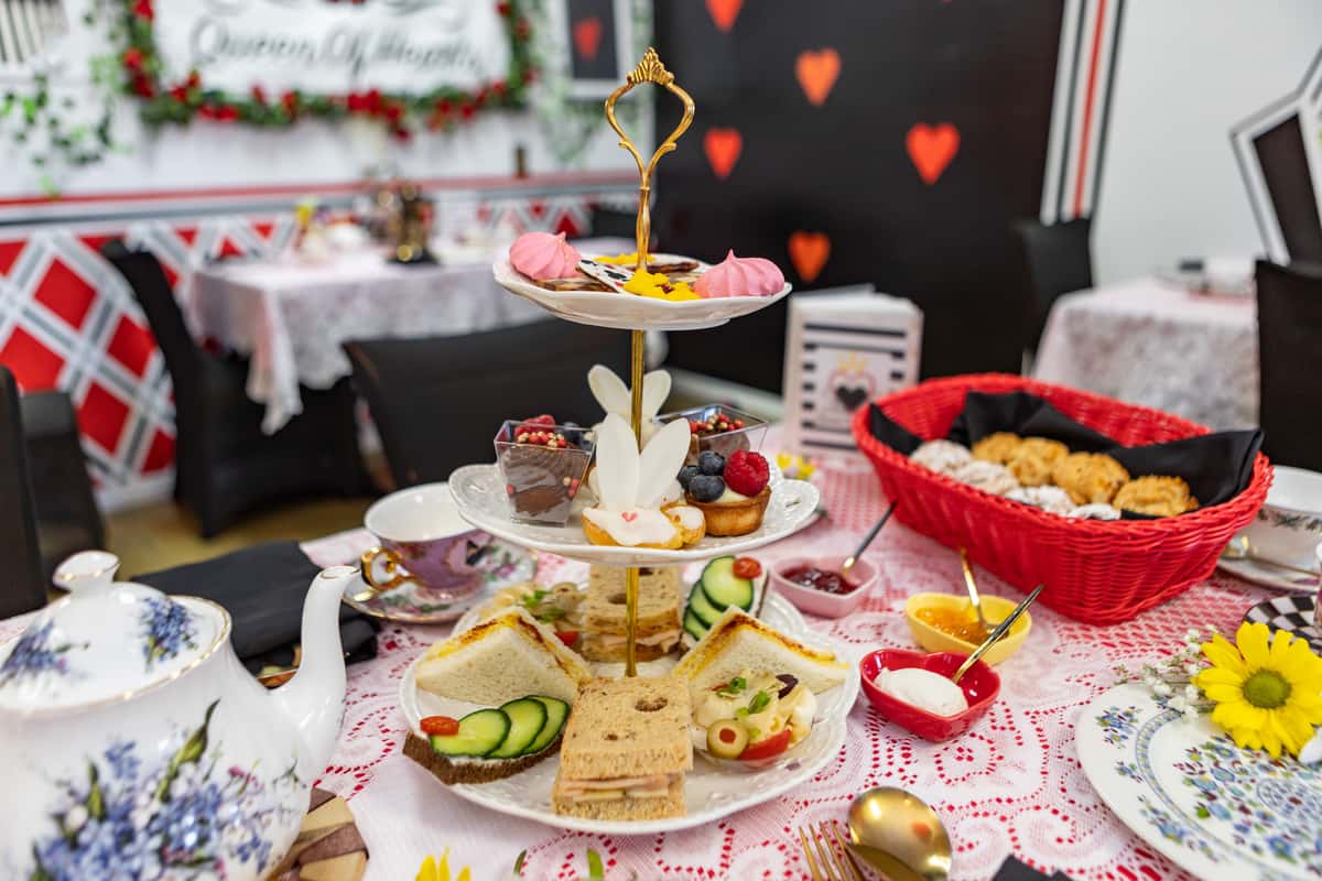 Alice in Wonderland-themed afternoon tea tower with finger sandwiches, sweets, scones, and floral china in a whimsical tea room.