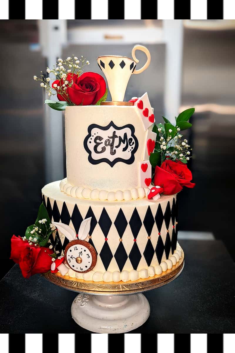 Alice in Wonderland themed cake with "Eat Me" sign, teacup topper, roses, hearts, and harlequin pattern decoration.