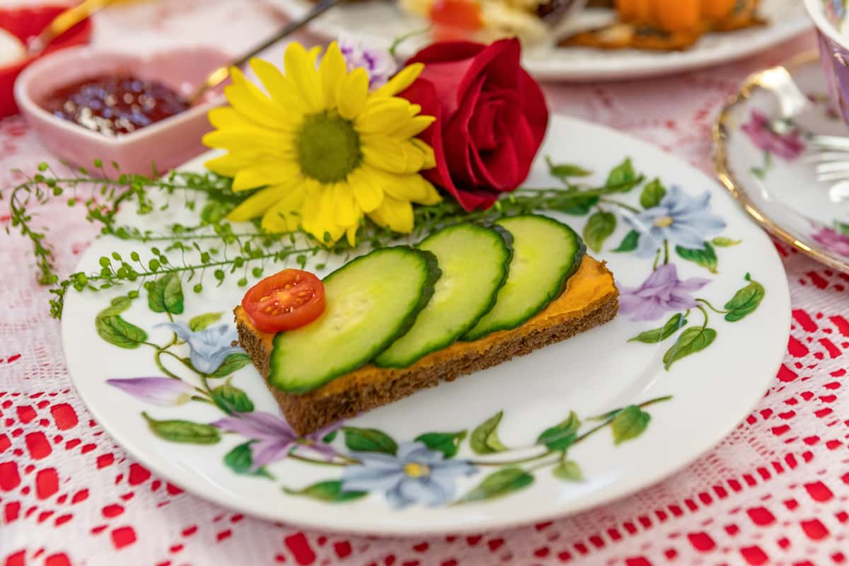 Open-faced cucumber sandwich on rye with a cherry tomato garnish, paired with floral decor on a vintage tea plate.
