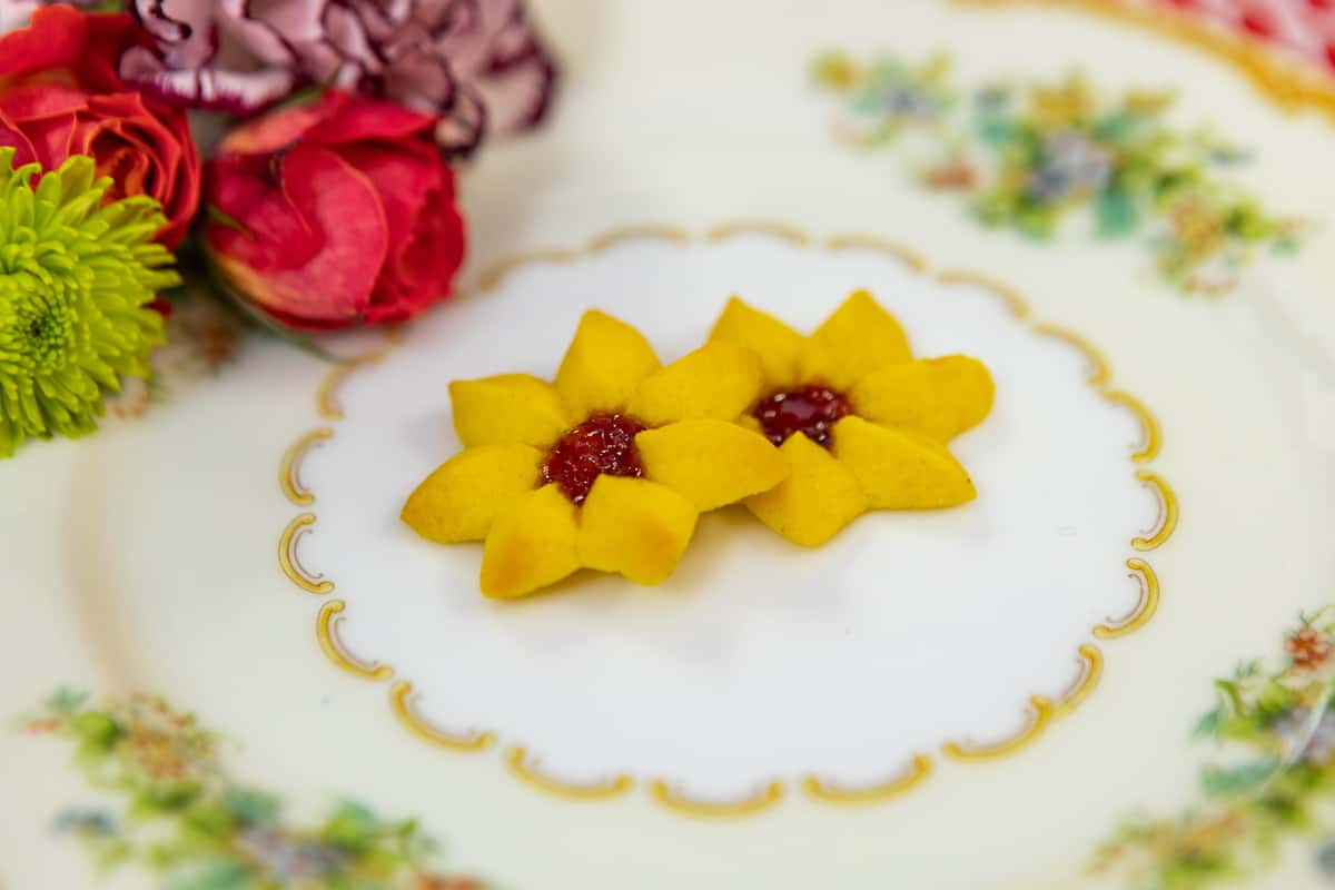 Sunflower-shaped shortbread cookies with a red jam center, served on ornate floral china with a garnish of fresh roses and mums.