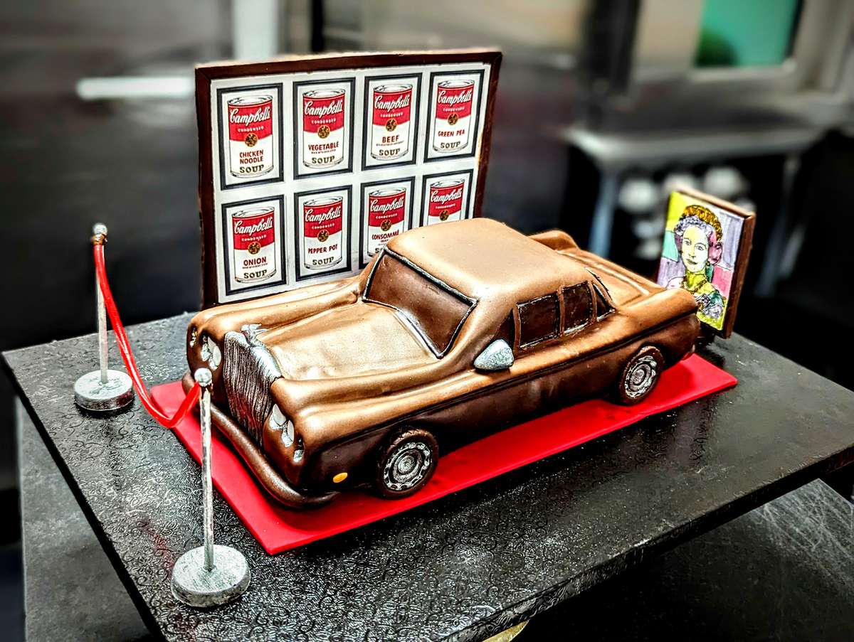 Sculpted cake shaped like a vintage car displayed with Andy Warhol style Campbell's soup art and pop art themed decorations.