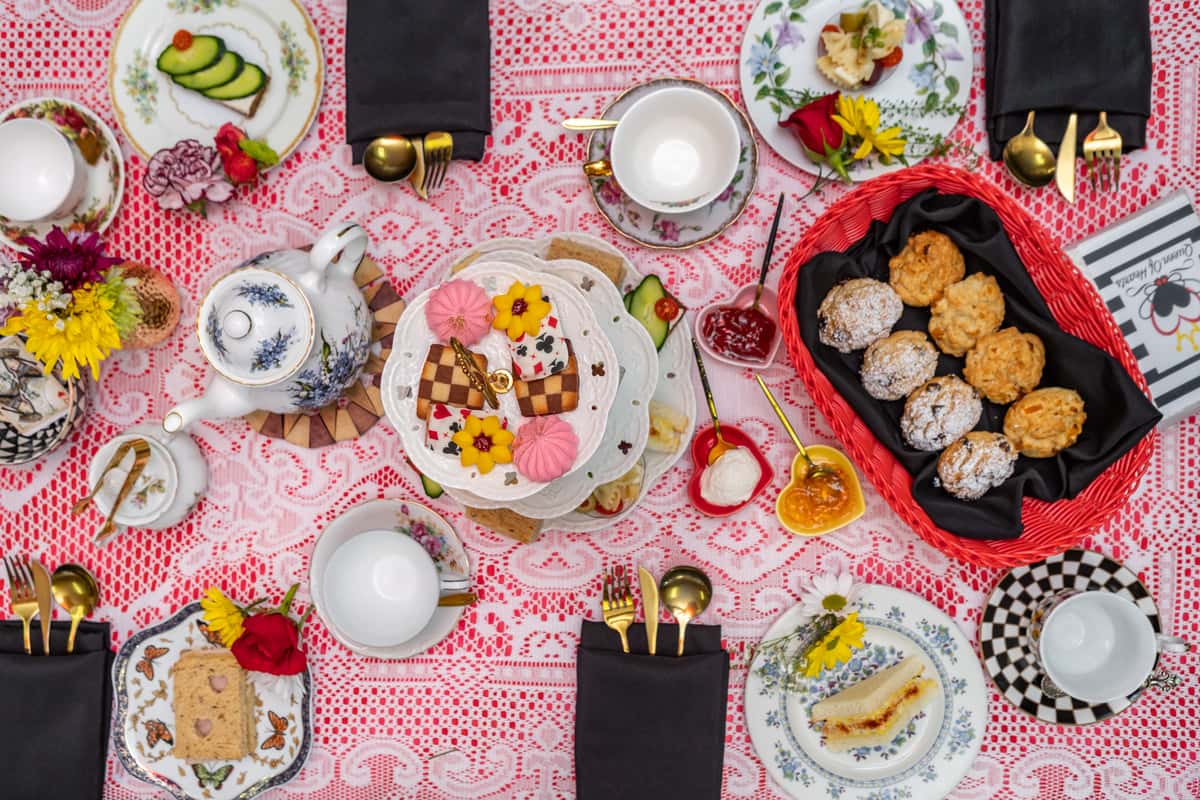 Overhead view of a whimsical afternoon tea spread featuring scones, tea sandwiches, cookies, and floral china on a lacy red tablecloth.